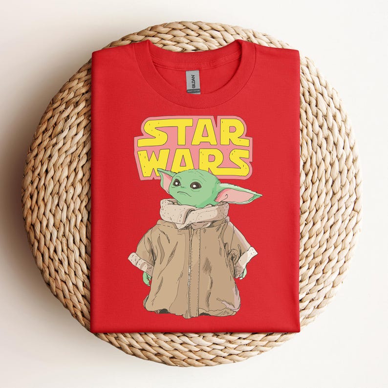 Peut inclure: T-shirt rouge avec un motif d'un personnage extraterrestre vert de Star Wars, avec le texte "STAR WARS" en jaune et rose. L'extraterrestre porte un manteau marron. Le t-shirt est pr&eacute;sent&eacute; sur une surface tiss&eacute;e.