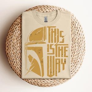May include: A beige t-shirt with a distressed gold graphic of a helmet and the text "THIS IS THE WAY." The shirt is folded on a round woven placemat. The shirt is made of a soft material.