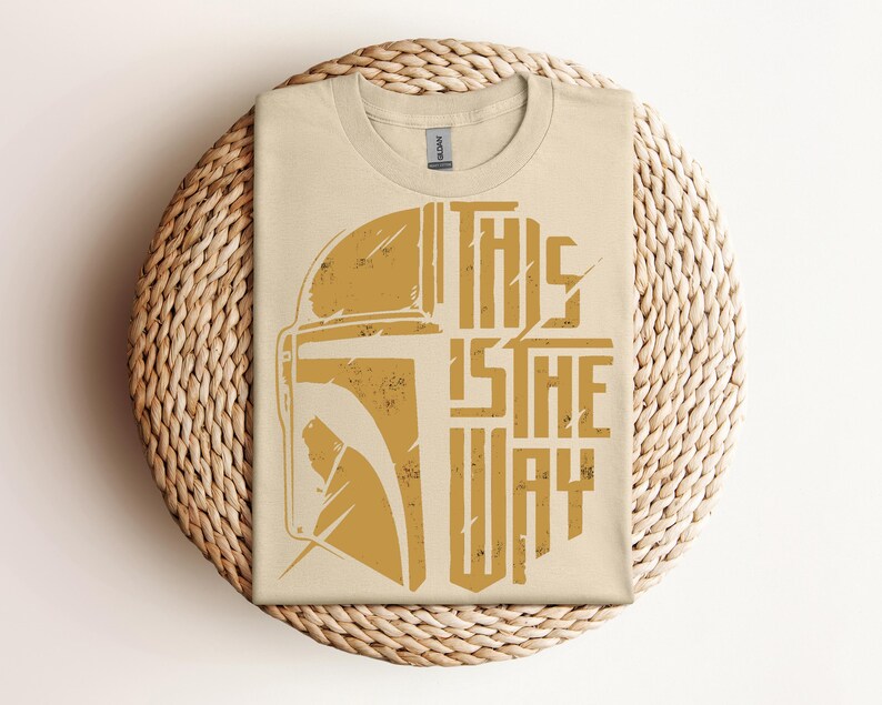 May include: A beige t-shirt with a distressed gold graphic of a helmet and the text "THIS IS THE WAY." The shirt is folded on a round woven placemat. The shirt is made of a soft material.