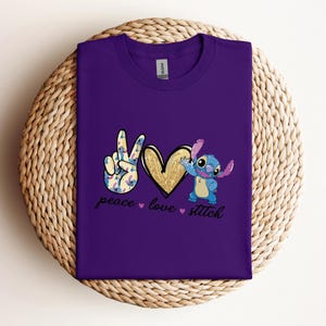以下が含まれることがあります： リロ＆スティッチのスティッチが描かれた紫色のTシャツ。平和のサイン、ハート、そして「peace love stitch」の文字が書かれています。