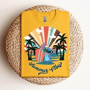 May include: Mustard yellow t-shirt with a cartoon character wearing sunglasses and holding a surfboard. The design shows a beach scene with palm trees, a sunset, and the words "Summer Vibes" in blue script. The shirt is folded on a woven surface.