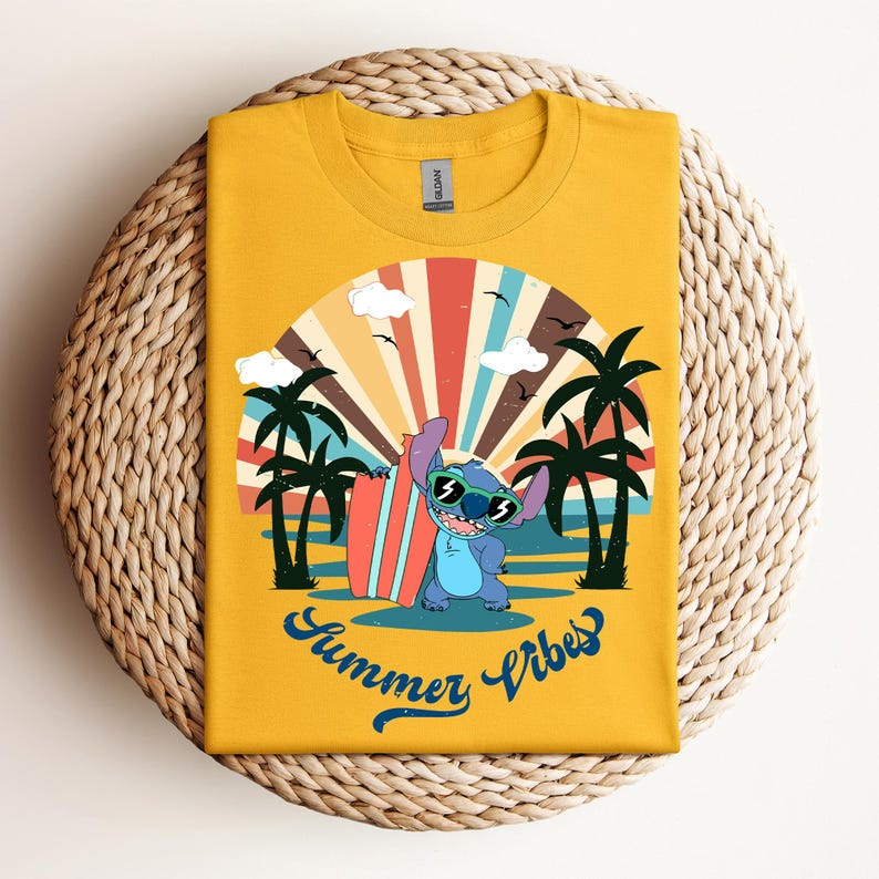 May include: Mustard yellow t-shirt with a cartoon character wearing sunglasses and holding a surfboard. The design shows a beach scene with palm trees, a sunset, and the words "Summer Vibes" in blue script. The shirt is folded on a woven surface.