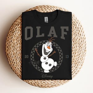 Może przedstawiać: Czarny T-shirt z białym nadrukiem Olafa z Krainy Lodu. Nadruk przedstawia Olafa w kwadratowym k&oacute;łku z tekstem "Olaf" nad nim i "Disney" pod nim.