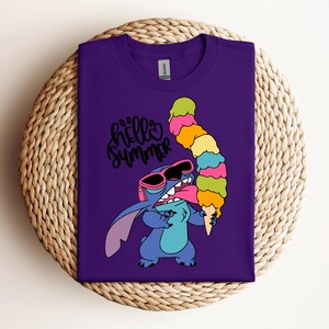 May include: A purple t-shirt featuring a cartoon character wearing sunglasses and holding a large, colorful ice cream cone. The words "hello summer" are written in a cursive font above the character. The shirt is displayed on a woven surface.