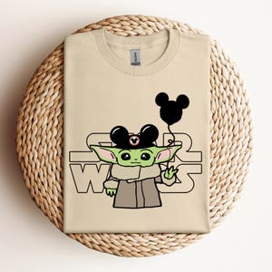 Puede incluir: Una camiseta beige con un personaje alien&iacute;gena verde que lleva un sombrero de Mickey Mouse y sostiene un globo negro. El texto "Star Wars" est&aacute; impreso en la camiseta.