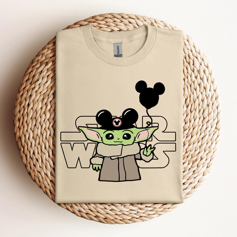 Puede incluir: Una camiseta beige con un personaje alien&iacute;gena verde que lleva un sombrero de Mickey Mouse y sostiene un globo negro. El texto "Star Wars" est&aacute; impreso en la camiseta.