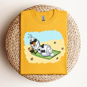 Op de afbeelding: Een geel T-shirt met een cartoon afbeelding van Olaf uit Frozen, die een zonnebril draagt en ontspant op een strandhanddoek met een kokosnootdrankje. De tekst "Let it Go" staat op het shirt gedrukt.