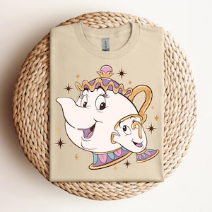 Könnte beinhalten: Beiges T-Shirt mit einer Cartoon-Illustration von Madame Pottine und Tassilo aus Die Schöne und das Biest. Das Design zeigt eine lächelnde Teekanne und Tasse mit goldenen, lila und rosa Akzenten und Sternen.