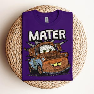 May include: A purple t-shirt featuring a cartoon image of Mater from the movie Cars. The word "MATER" is printed above the character in white block letters. The graphic is in shades of orange, blue, and brown.