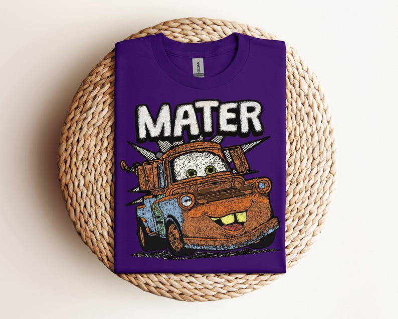 May include: A purple t-shirt featuring a cartoon image of Mater from the movie Cars. The word "MATER" is printed above the character in white block letters. The graphic is in shades of orange, blue, and brown.