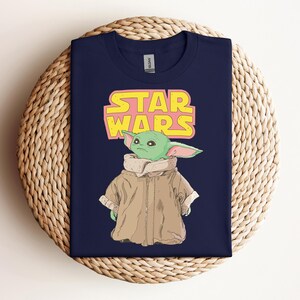 Peut inclure: T-shirt bleu marine avec un dessin d'un personnage extraterrestre vert portant un manteau beige. Le logo Star Wars est au-dessus du personnage en jaune et rose. Le t-shirt est pr&eacute;sent&eacute; sur une surface tiss&eacute;e.