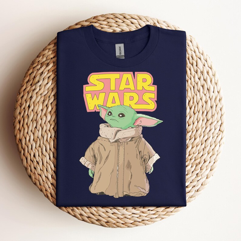 Peut inclure: T-shirt bleu marine avec un dessin d'un personnage extraterrestre vert portant un manteau beige. Le logo Star Wars est au-dessus du personnage en jaune et rose. Le t-shirt est pr&eacute;sent&eacute; sur une surface tiss&eacute;e.