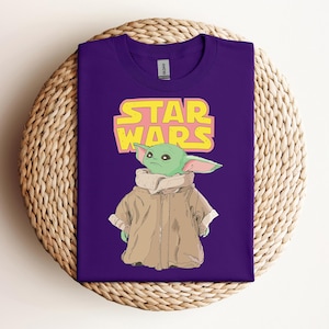 Peut inclure: Un t-shirt violet avec un graphisme d'un personnage extraterrestre vert de Star Wars, portant un manteau beige. Les mots "STAR WARS" sont imprim&eacute;s en jaune et rose au-dessus du personnage.