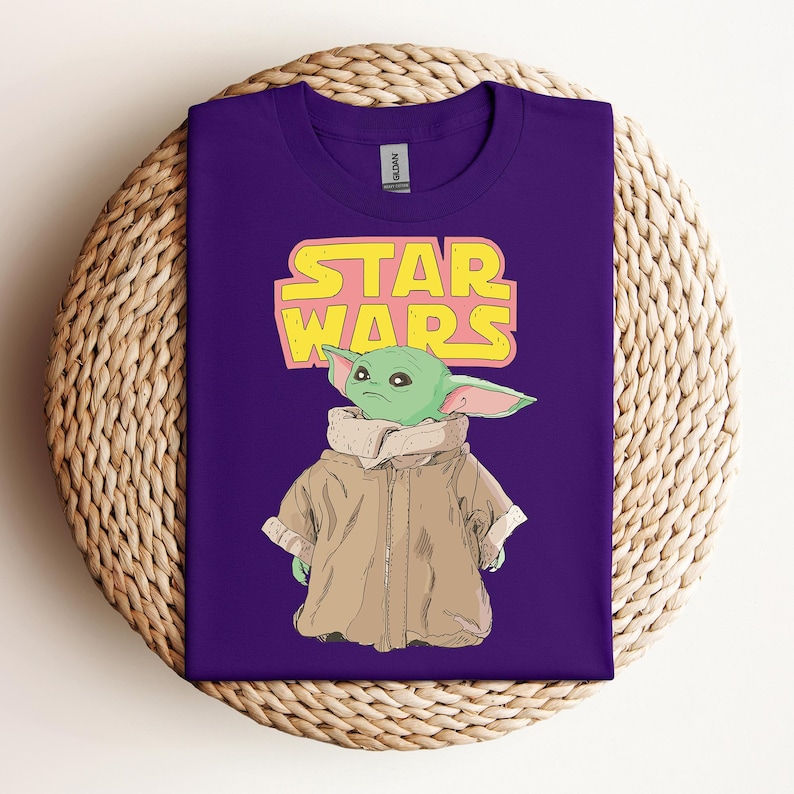 Peut inclure: Un t-shirt violet avec un graphisme d'un personnage extraterrestre vert de Star Wars, portant un manteau beige. Les mots "STAR WARS" sont imprim&eacute;s en jaune et rose au-dessus du personnage.
