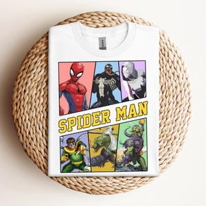 Pode incluir: Camiseta branca com um gr&aacute;fico de banda desenhada com o Homem-Aranha e outros personagens. O design inclui o texto "SPIDER MAN" em amarelo e preto. A camisa est&aacute; dobrada sobre uma superf&iacute;cie circular tecida.