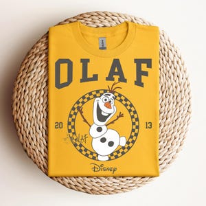 Może przedstawiać: Ż&oacute;łta koszulka z czarno-białym nadrukiem Olafa z Krainy Lodu. Nadruk zawiera tekst "Olaf" czarną czcionką w stylu uniwersyteckim i "Disney" mniejszą czarną czcionką. Nadruk zawiera r&oacute;wnież kwadratowe koło wok&oacute;ł Olafa z liczbami "20" i "13" po obu stronach.
