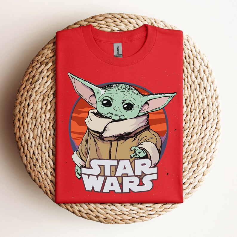 Op de afbeelding: Rood t-shirt met een afbeelding van Grogu uit Star Wars. Het ontwerp bevat Grogu in een cirkelvormige achtergrond, met de woorden "STAR WARS" in het wit eronder. Het shirt is gemaakt van een zacht materiaal.