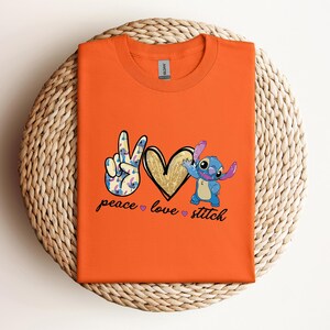 以下が含まれることがあります： リロ＆スティッチのスティッチが描かれたオレンジ色のTシャツ。平和のサイン、ハート、そして「peace love stitch」の文字が書かれています。