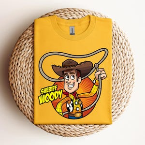 May include: Mustard yellow t-shirt featuring a cartoon illustration of Sheriff Woody from Toy Story. The design includes Woody in his cowboy hat, holding a lasso, with the text "Sheriff Woody" in a yellow and white font.
