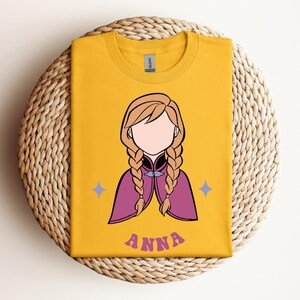 May include: A mustard yellow t-shirt featuring a graphic illustration of a character with blonde hair in two braids, wearing a magenta cape. The character has no facial features. Two small light purple stars flank the figure, and the name ANNA is printed below in a wavy, purple, retro-style font.