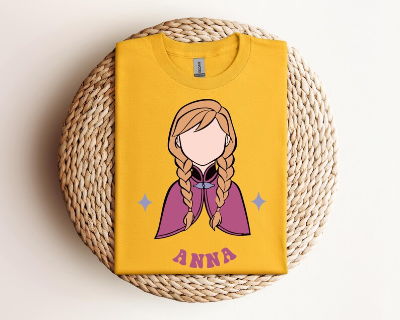 May include: A mustard yellow t-shirt featuring a graphic illustration of a character with blonde hair in two braids, wearing a magenta cape. The character has no facial features. Two small light purple stars flank the figure, and the name ANNA is printed below in a wavy, purple, retro-style font.