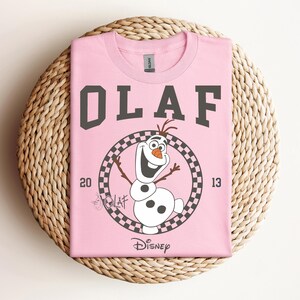 Może przedstawiać: R&oacute;żowa koszulka z czarno-białym kwadratowym kołem przedstawiającym Olafa z Krainy Lodu. Tekst "OLAF" jest czarny i znajduje się nad kołem, a "DISNEY" pod kołem.