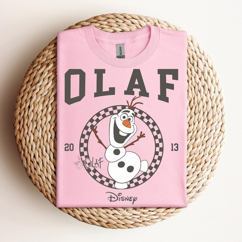 Może przedstawiać: R&oacute;żowa koszulka z czarno-białym kwadratowym kołem przedstawiającym Olafa z Krainy Lodu. Tekst "OLAF" jest czarny i znajduje się nad kołem, a "DISNEY" pod kołem.