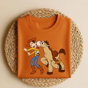 Peut inclure: Un t-shirt orange &agrave; col rond avec une illustration de dessin anim&eacute; de Woody et Bullseye de Toy Story. Woody porte un chapeau de cowboy, une chemise &agrave; carreaux jaune, un jean bleu et des bottes marron. Bullseye est un cheval brun avec une selle.
