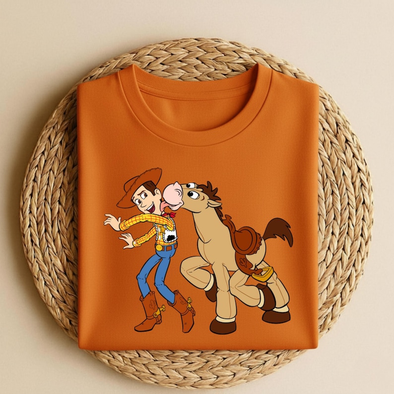 Peut inclure: Un t-shirt orange &agrave; col rond avec une illustration de dessin anim&eacute; de Woody et Bullseye de Toy Story. Woody porte un chapeau de cowboy, une chemise &agrave; carreaux jaune, un jean bleu et des bottes marron. Bullseye est un cheval brun avec une selle.
