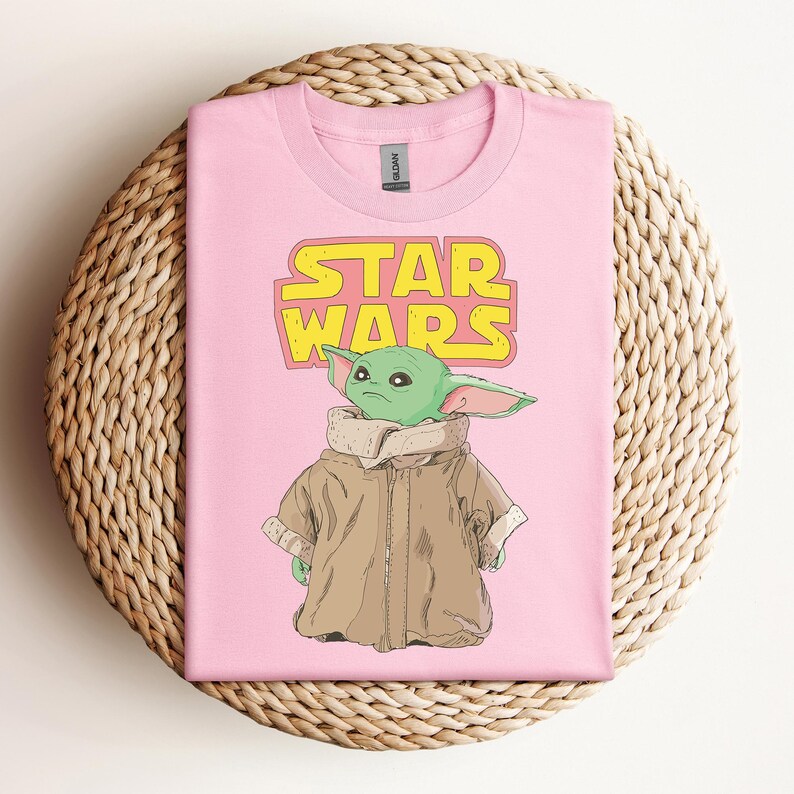 Peut inclure: T-shirt rose avec un graphisme d'un personnage extraterrestre vert de Star Wars, avec le logo Star Wars en jaune et orange. L'extraterrestre porte un manteau marron. Le t-shirt est pr&eacute;sent&eacute; sur une surface tiss&eacute;e.