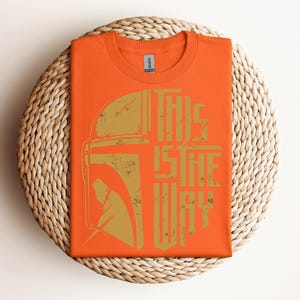 May include: Orange t-shirt with a gold graphic of a helmet and the text "THIS IS THE WAY." The shirt is folded on a woven, circular placemat. The design has a distressed, vintage look.