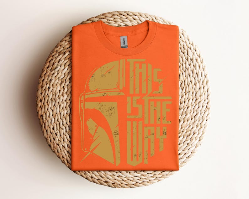 May include: Orange t-shirt with a gold graphic of a helmet and the text "THIS IS THE WAY." The shirt is folded on a woven, circular placemat. The design has a distressed, vintage look.