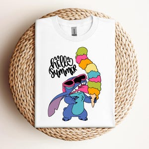 May include: White t-shirt featuring a cartoon character wearing sunglasses and holding a large, colorful ice cream cone. The shirt has the words "hello summer" in a playful font. The design is centered on the shirt.