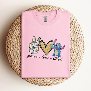 以下が含まれることがあります： リロ＆スティッチのスティッチが描かれたピンクのTシャツ。平和のサイン、ハート、そして「peace love stitch」の文字が書かれています。