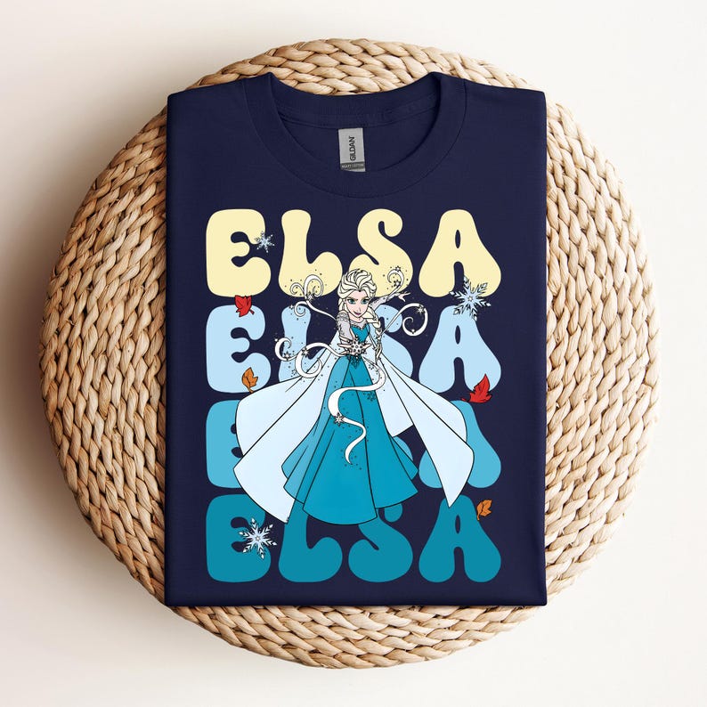 Op de afbeelding: Marineblauw T-shirt met het woord "ELSA" in een retro lettertype en een afbeelding van Elsa uit Frozen. Het ontwerp bevat sneeuwvlokken, bladeren en een portret van Elsa in een blauwe jurk en witte cape.