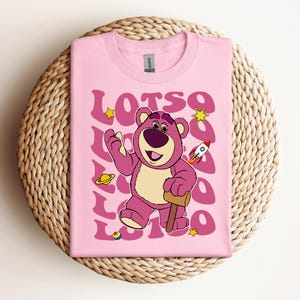 Puede incluir: Camiseta rosa con una imagen de dibujos animados de Lotso el oso de la franquicia Toy Story. El oso es rosa con una estrella amarilla y un cohete en el fondo. El texto "LOTSO" se repite en una fuente retro.