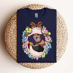 Puede incluir: Camiseta azul marino con un personaje de dibujos animados dentro de un marco ovalado floral. El personaje es morado y marr&oacute;n, rodeado de flores rosas, amarillas y azules, y una cinta marr&oacute;n. El dise&ntilde;o est&aacute; centrado en la camiseta.