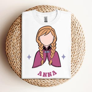 May include: A white t-shirt featuring a graphic illustration of a character with blonde braided hair, wearing a magenta cape with a dark collar. The design includes two small light purple four-pointed stars and the name ANNA in pink letters.