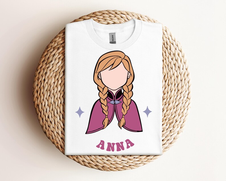 May include: A white t-shirt featuring a graphic illustration of a character with blonde braided hair, wearing a magenta cape with a dark collar. The design includes two small light purple four-pointed stars and the name ANNA in pink letters.