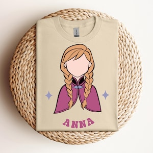 May include: A beige t-shirt featuring a graphic illustration of a character with blonde braided hair, wearing a magenta cape with a dark collar. The design includes two small light purple four-pointed stars and the name ANNA in pink letters.