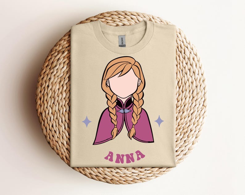 May include: A beige t-shirt featuring a graphic illustration of a character with blonde braided hair, wearing a magenta cape with a dark collar. The design includes two small light purple four-pointed stars and the name ANNA in pink letters.