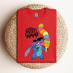 May include: Red t-shirt featuring a cartoon character wearing sunglasses and holding a large, colorful ice cream cone. The text "hello summer" is written in black script. The shirt is displayed on a woven, circular surface.