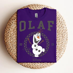 Może przedstawiać: Fioletowy T-shirt z grafiką Olafa z Krainy Lodu. Grafika to koło z szachownicą i tekstem "Olaf" dużą czcionką nad kołem. Tekst "20" i "13" znajduje się po obu stronach koła. Tekst "Disney" znajduje się pod kołem.