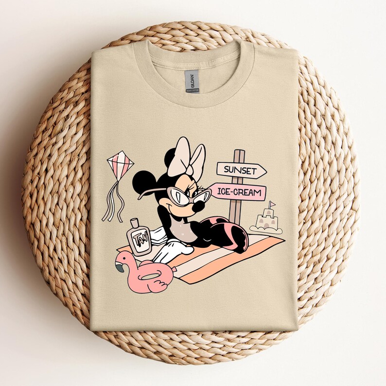 K&ouml;nnte beinhalten: Ein beiges T-Shirt mit einem Cartoon-Bild von Minnie Mouse, die eine Sonnenbrille und einen Badeanzug tr&auml;gt. Sie sitzt auf einem Strandtuch mit einem rosafarbenen Flamingo-Schwimmring, einer Flasche Sonnencreme und einem Schild mit der Aufschrift "Sunset" und "Ice-Cream".