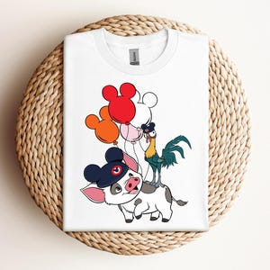 Op de afbeelding: Wit T-shirt met een cartoon illustratie van een varken met een marineblauwe hoed, een haan en Mickey Mouse-vormige ballonnen. Het T-shirt is opgevouwen op een ronde, geweven ondergrond.