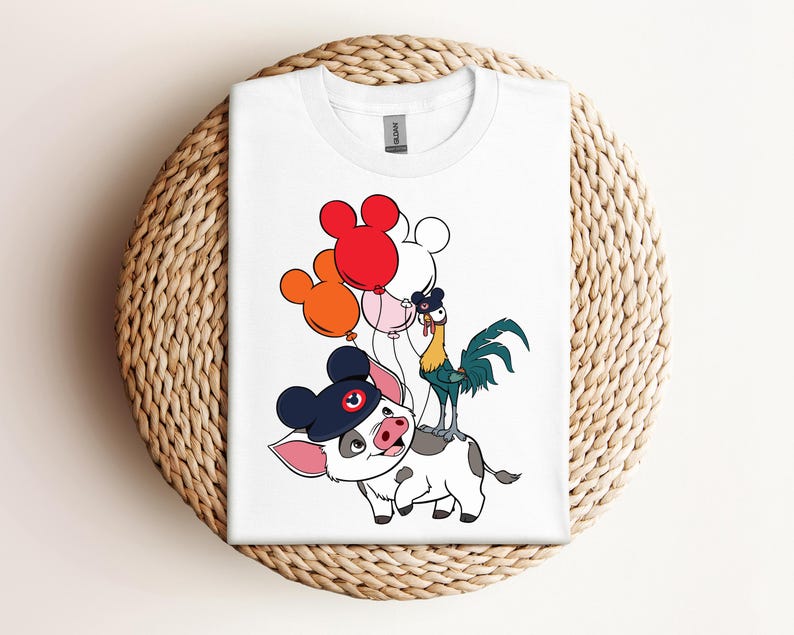 Op de afbeelding: Wit T-shirt met een cartoon illustratie van een varken met een marineblauwe hoed, een haan en Mickey Mouse-vormige ballonnen. Het T-shirt is opgevouwen op een ronde, geweven ondergrond.