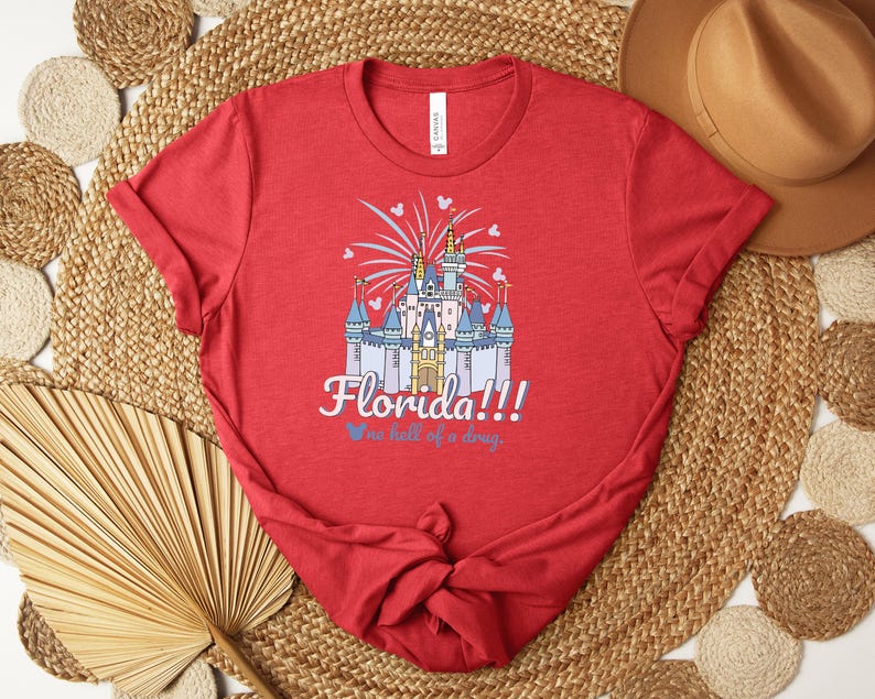 Puede incluir: Camiseta roja con un castillo y la palabra "Florida!!!" en escritura blanca. La frase "one hell of a day" est&aacute; debajo en azul. La camiseta est&aacute; anudada en la parte inferior. La camiseta es de algod&oacute;n.