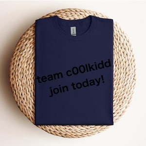 May include: Navy blue t-shirt with the text "team c00lkidd join today!" in black. The shirt is folded and placed on a round, woven wicker placemat. The Gildan brand label is visible at the neckline.