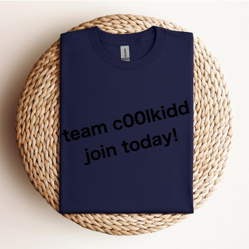 May include: Navy blue t-shirt with the text "team c00lkidd join today!" in black. The shirt is folded and placed on a round, woven wicker placemat. The Gildan brand label is visible at the neckline.