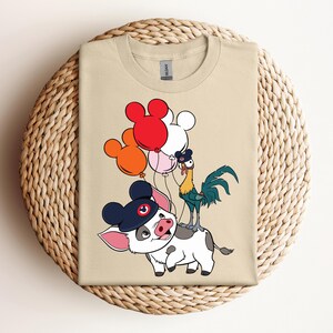 Op de afbeelding: Beige T-shirt met een cartoonvarken met een marineblauwe hoed met een Mickey Mouse-ontwerp. Een haan en kleurrijke ballonnen zijn ook aanwezig. Het shirt ligt op een geweven, cirkelvormig oppervlak.